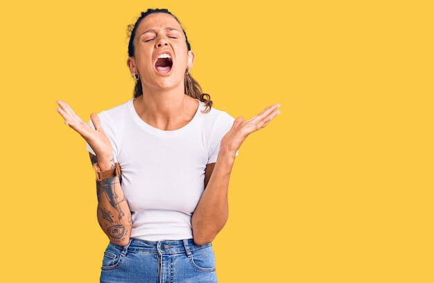 Young hispanic woman with tattoo wearing casual white tshirt celebrating mad and crazy for success with arms raised and closed eyes screaming excited winner concept