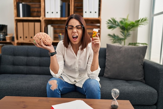 Young hispanic woman working on depression holding pills angry and mad screaming frustrated and furious, shouting with anger. rage and aggressive concept.