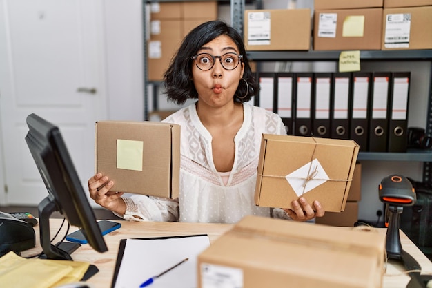 Young hispanic woman working at small business ecommerce making fish face with mouth and squinting eyes, crazy and comical.