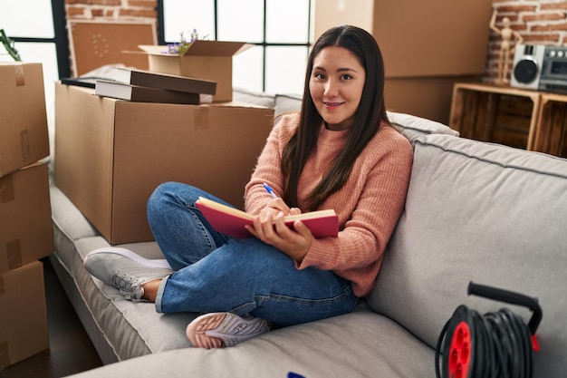 Young hispanic woman writing on book sitting on sofa at new home
