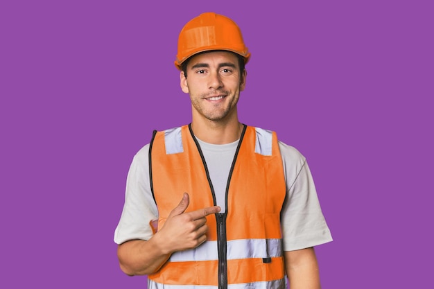 Young Hispanic worker in safety gear person pointing by hand to a shirt copy space proud