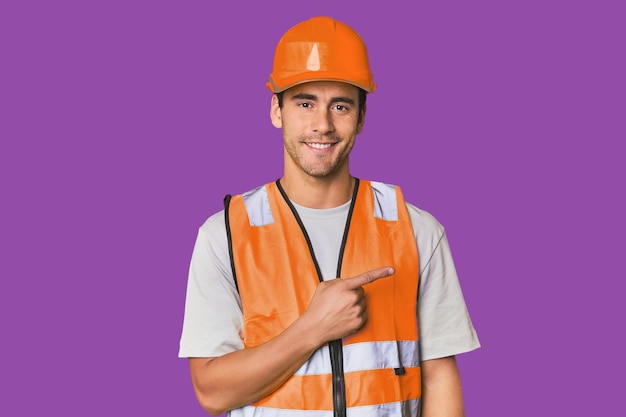 Young Hispanic worker in safety gear smiling and pointing aside showing something at blank space