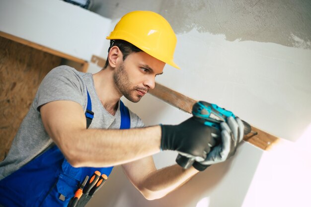 Foto giovane maschio riparatore domestico o lavoratore impegnato con la ristrutturazione della casa, tiene in mano attrezzature per l'edilizia, indossa abiti da lavoro casuali nella nuova costruzione