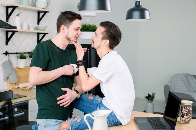 Young homosexual lovers looking at one another at home. Happy smiling gay couple drinking coffee together at kitchen