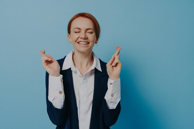 Young hopeful businesswoman with closed eyes in formal wear crossing fingers isolated on blue