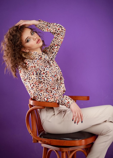 Young hot curlyhaired girl sitting on a chair and looking at he camera on purple background High quality photo