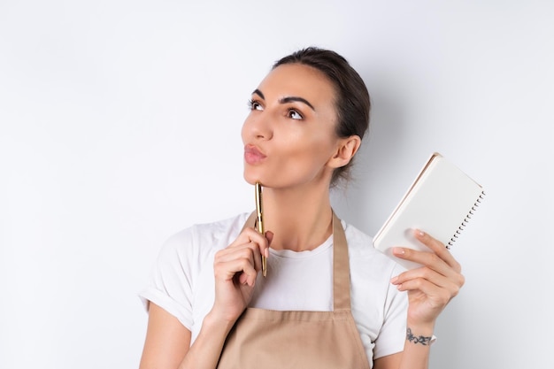 A young housewife in an apron on a white background with a notebook in her hands chooses recipes for dinner thinks how to please her husband comes up with ideas for dishes