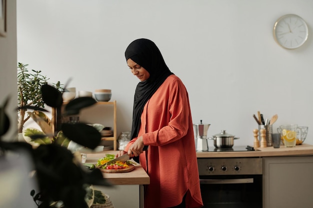 Young housewife in red shirt and black hijab cutting fresh vegetables on chopping board