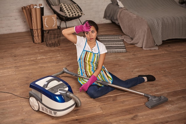 A young housewife sitting leaning on vacuum cleaner tired of housework.