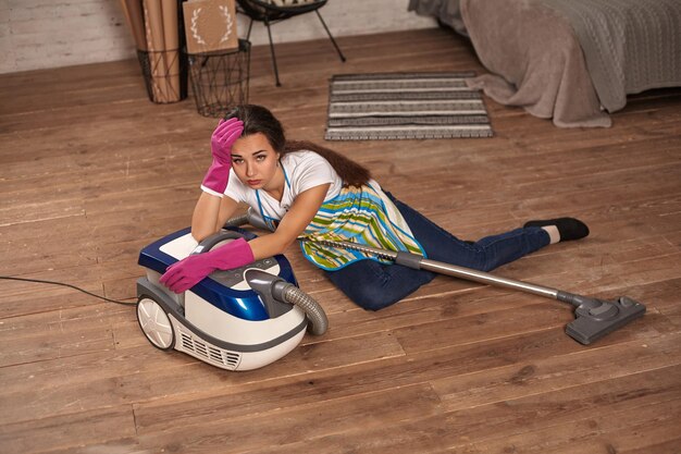 A young housewife sitting leaning on vacuum cleaner tired of housework.