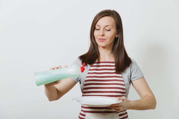 Giovane casalinga in grembiule a strisce isolato. la donna della governante tiene la bottiglia con un liquido più pulito per lavare i piatti, piatto rotondo vuoto bianco