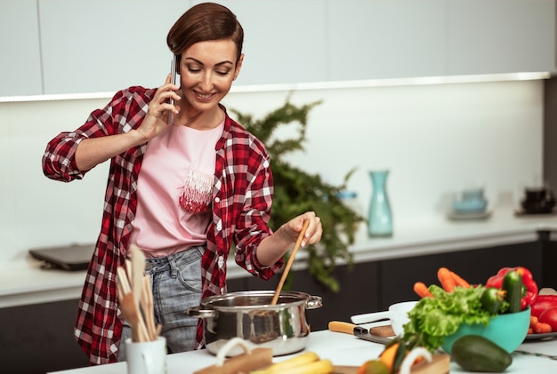 Young housewife talking on the phone while mixing a sup in a pan with a short hairstyle while