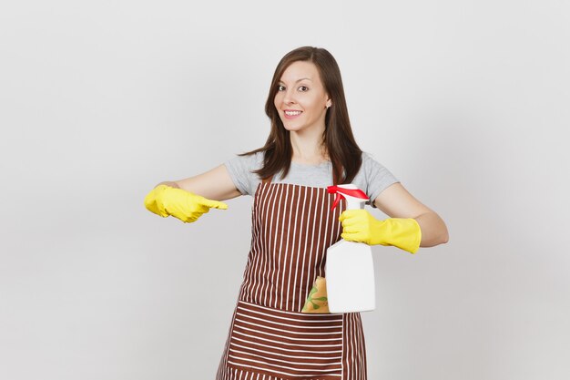 Young housewife in yellow gloves, striped apron, cleaning rag in pocket isolated on white background. Woman holding and pointing on bottle with cleaner liquid, sponge. Copy space for advertisement.