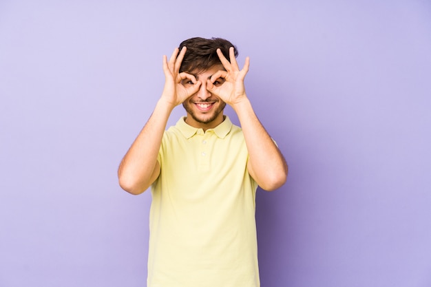 Young ian man on a purple wall showing okay sign over eyes