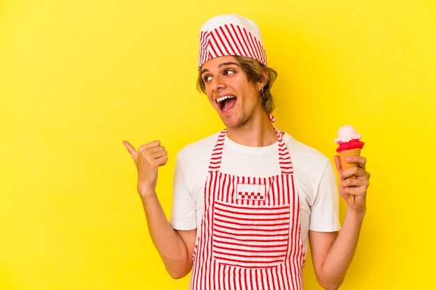 Молодой человек производителя мороженого с косметикой, держащей мороженое, изолированное на желтом фоне, указывает пальцем далеко, смеясь и беззаботно.