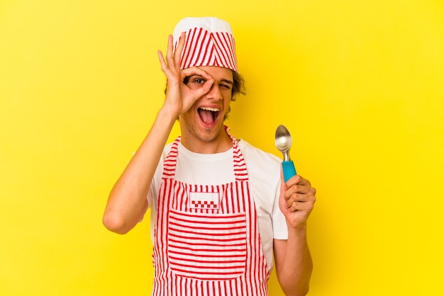 Young ice cream maker man with makeup holding spoon isolated on yellow background  excited keeping ok gesture on eye.