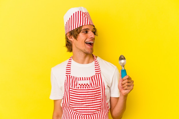 Young ice cream maker man with makeup holding spoon isolated on yellow background  looks aside smiling, cheerful and pleasant.