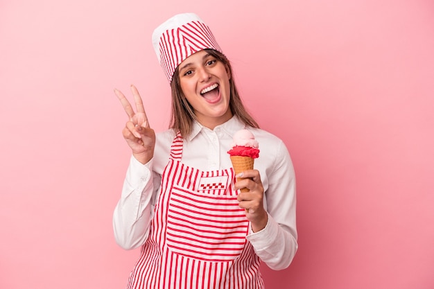 young ice cream maker woman holding ice cream isolated on pink background joyful and carefree showing a peace symbol with fingers.