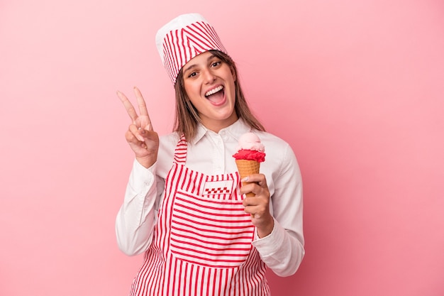 Young ice cream maker woman holding ice cream isolated on pink background joyful and carefree showing a peace symbol with fingers.