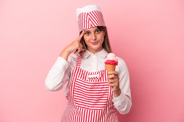 Young ice cream maker woman holding ice cream isolated on pink background pointing temple with finger, thinking, focused on a task.