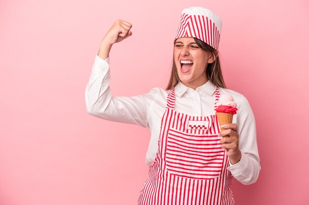 young ice cream maker woman holding ice cream isolated on pink background raising fist after a victory, winner concept.