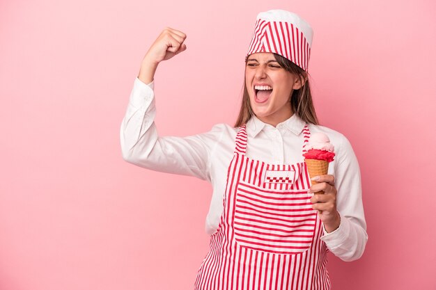 Young ice cream maker woman holding ice cream isolated on pink background raising fist after a victory, winner concept.