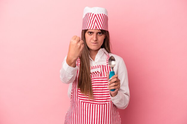 カメラに拳、攻撃的な表情を示すピンクの背景に分離されたスプーンを保持している若いアイスクリームメーカーの女性。