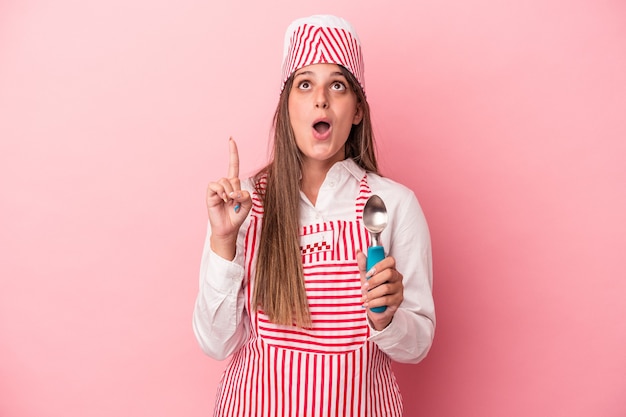 Young ice cream maker woman holding spoon isolated on pink background pointing upside with opened mouth.