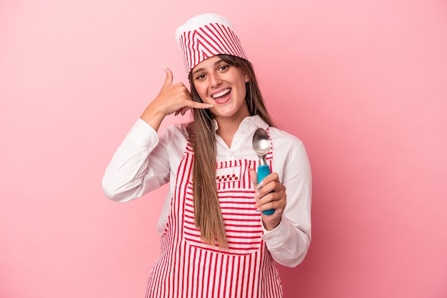 Giovane gelatiera donna che tiene il cucchiaio isolato su sfondo rosa che mostra un gesto di chiamata del telefono cellulare con le dita.