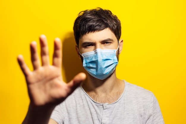 young ill man wearing medical flu mask, shows gesture STOP with hand on wall of yellow.