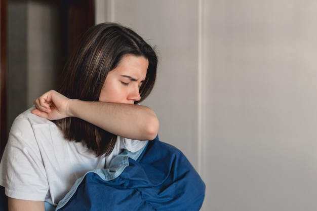 Young ill woman infected by the coronavirus is coughing into her upper sleeve while covering with a blue quilt in her home