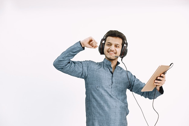 Young Indaian guy in headphones listening to music. Dancing on white wall, isolated.