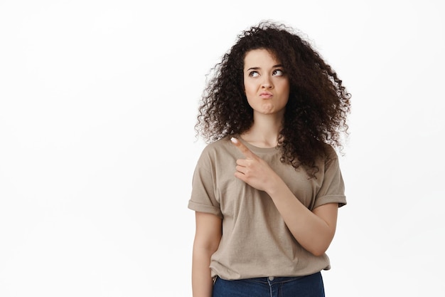 Young indecisive woman pointing and looking at upper left corner, making perplexed face expression, white background