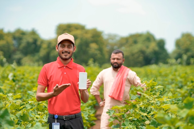 Молодой индийский агроном или банкир показывает смартфон с фермерами на сельскохозяйственном поле.