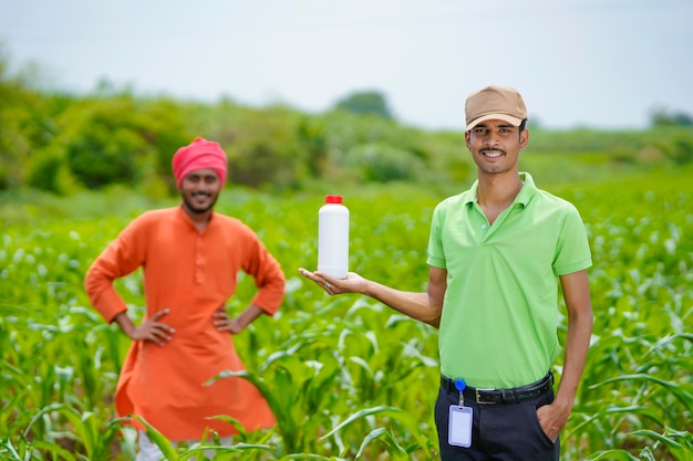 Young indian agronomist holding liquid fertilizer bottle with farmer at green agriculture field.