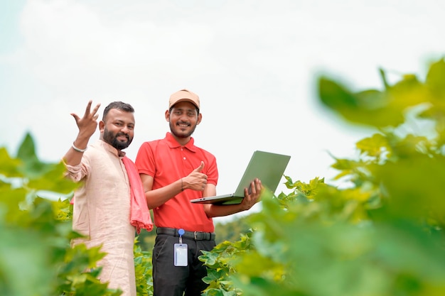 Young indian agronomist showing information to farmer in laptop at green agriculture field.