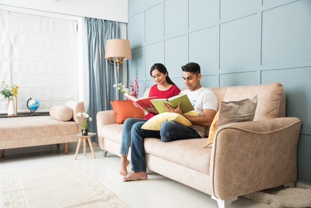 Young Indian asian couple reading books while sitting on sofa at home, modern living space