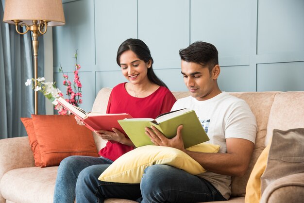 Young Indian asian couple reading books while sitting on sofa at home, modern living space