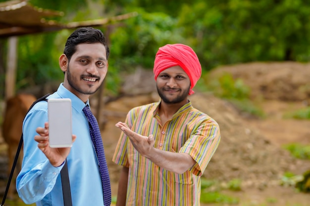 Giovane funzionario di banca indiano o agronomo che mostra smartphone con un agricoltore nella sua fattoria