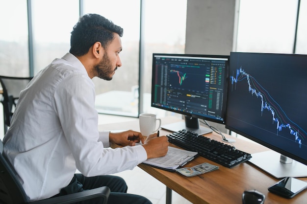 Young indian business man trader looking at computer screen with trading charts reflecting in eyeglasses watching stock trading market financial data growth concept close up