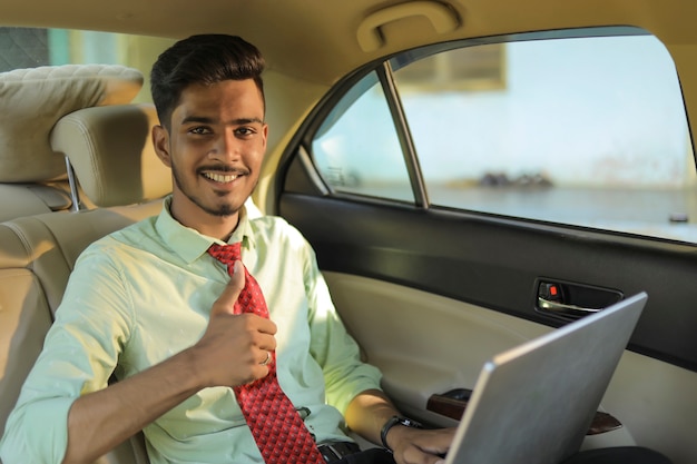 Молодой индийский деловой человек, использующий ноутбук и показывающий бугорки в машине