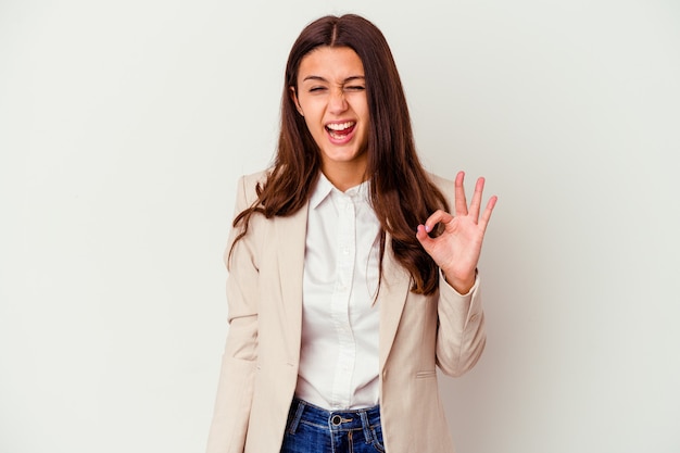 Young Indian business woman isolated on white winks an eye and holds an okay gesture with hand.