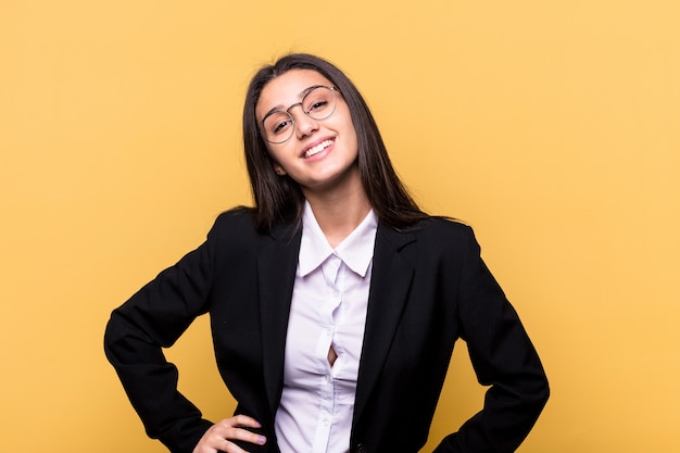 Young Indian business woman isolated on yellow wall confident keeping hands on hips.