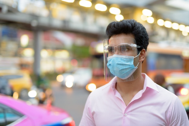 Молодой индийский бизнесмен с маской и щитком для лица думает на улицах города