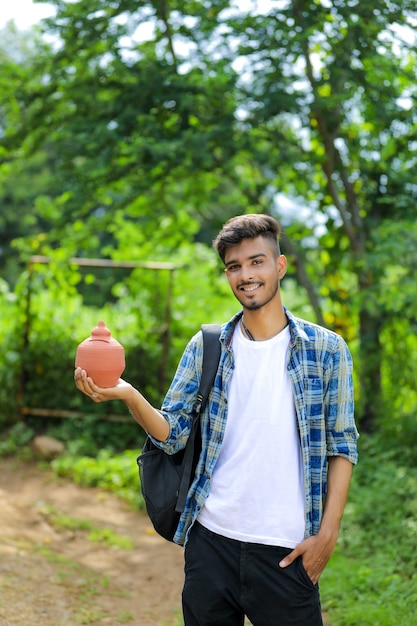 Young indian college boy holding clay piggy bank in hand over nature background