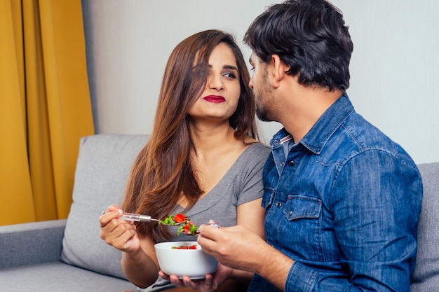 Young indian couple sitting in they living room and eating vegetable salad