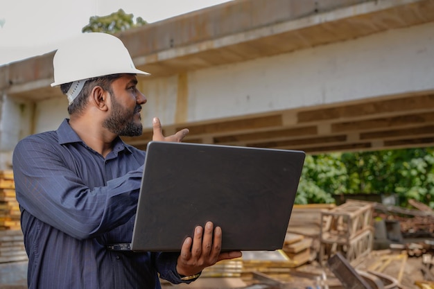 Young indian engineer in uniform and using laptop at field