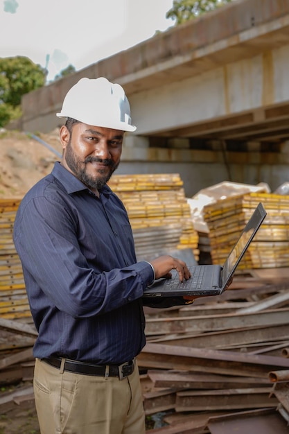 Young Indian engineer in uniform and using laptop at field