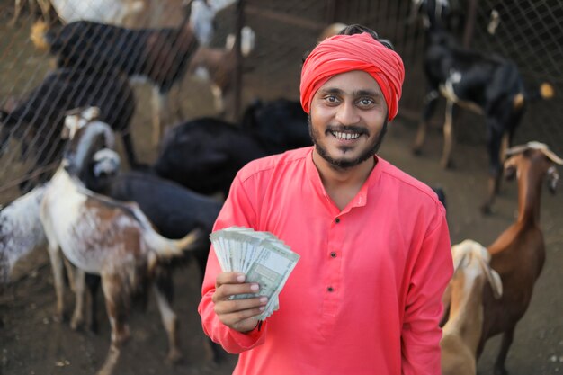 Young indian farmer showing money at goat dairy farm