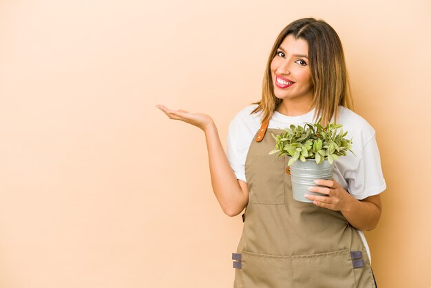 手のひらにコピースペースを示し、腰に別の手を保持している孤立した植物を保持している若いインドの庭師の女性